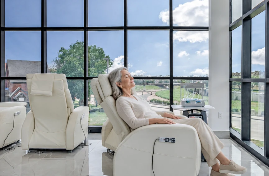 A client relaxing with her eyes closed in a reclining chair beside floor-to-ceiling windows at HealthCo